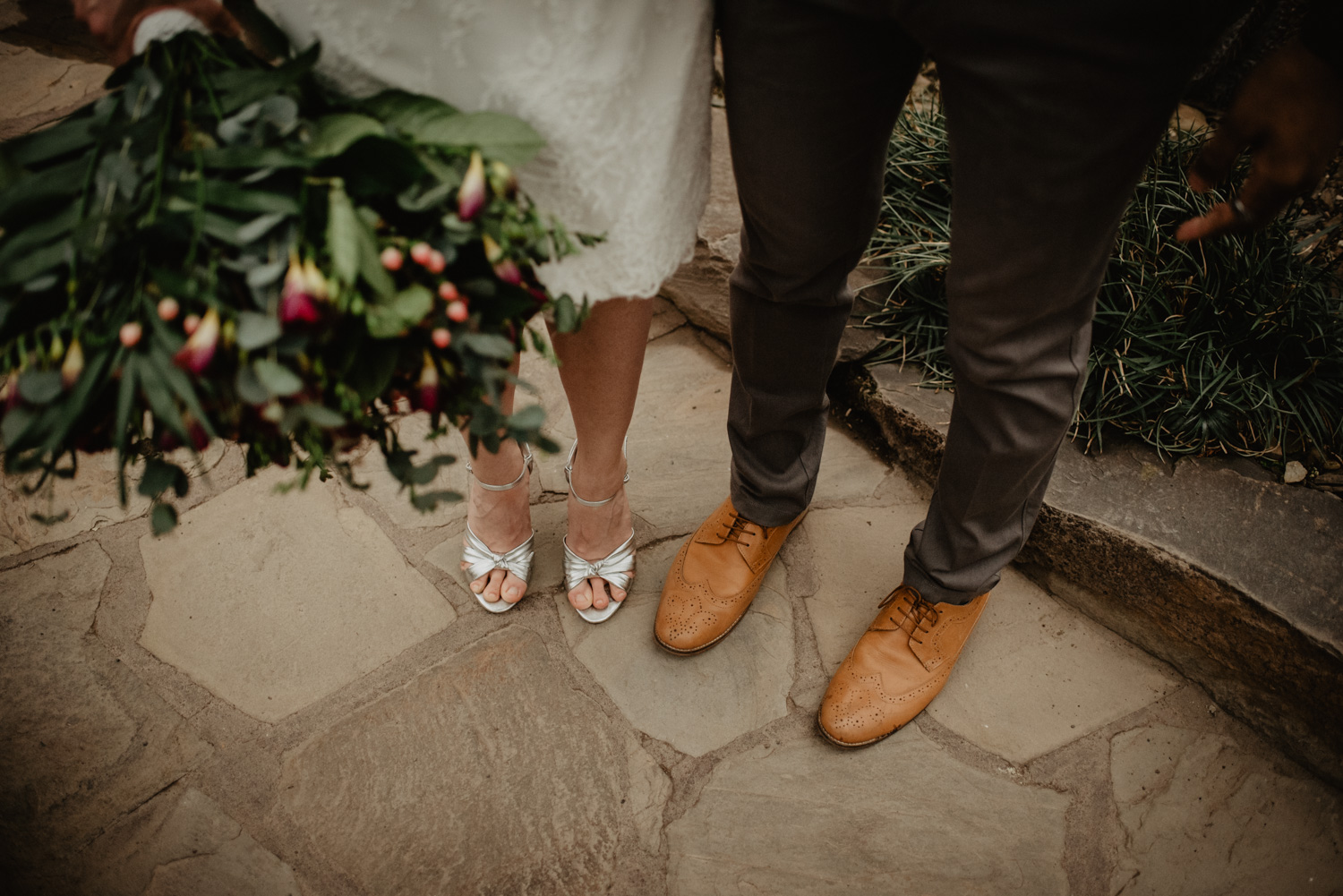 Detailfoto der Füße des Brautpaares. Sie hat ein kurzes Kleid und silberfarbene High Heels an, er eine dunkelgraue Stoffhose und hellbraune Anzugschuhe.