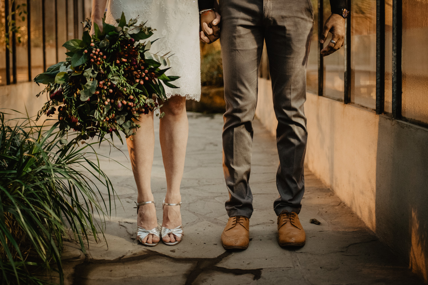 Detailfoto der Füße des Brautpaares. Sie hat ein kurzes Kleid und silberfarbene High Heels an, er eine dunkelgraue Stoffhose und hellbraune Anzugschuhe.