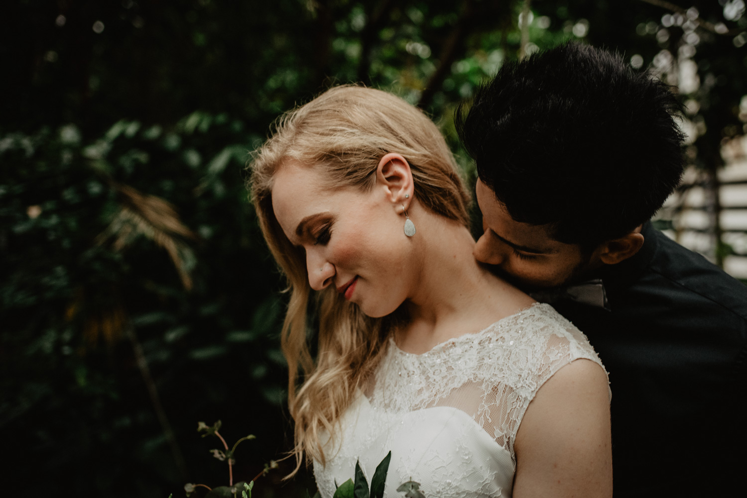 Ein Mann steht hinter seiner Braut und küsst sie am Hals. Sie hält einen Blumenstrauß und ihre langen blonden lockigen Haare liegen auf einer Seite.