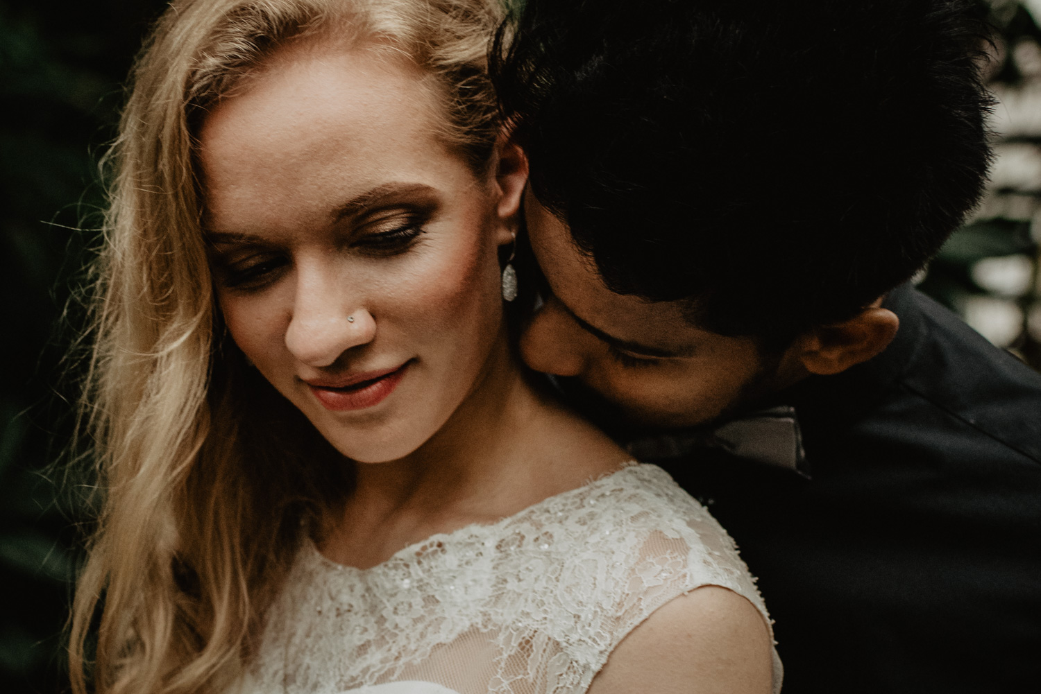 Ein Mann steht hinter seiner Braut und küsst sie am Hals. Ihre langen blonden lockigen Haare liegen auf einer Seite.