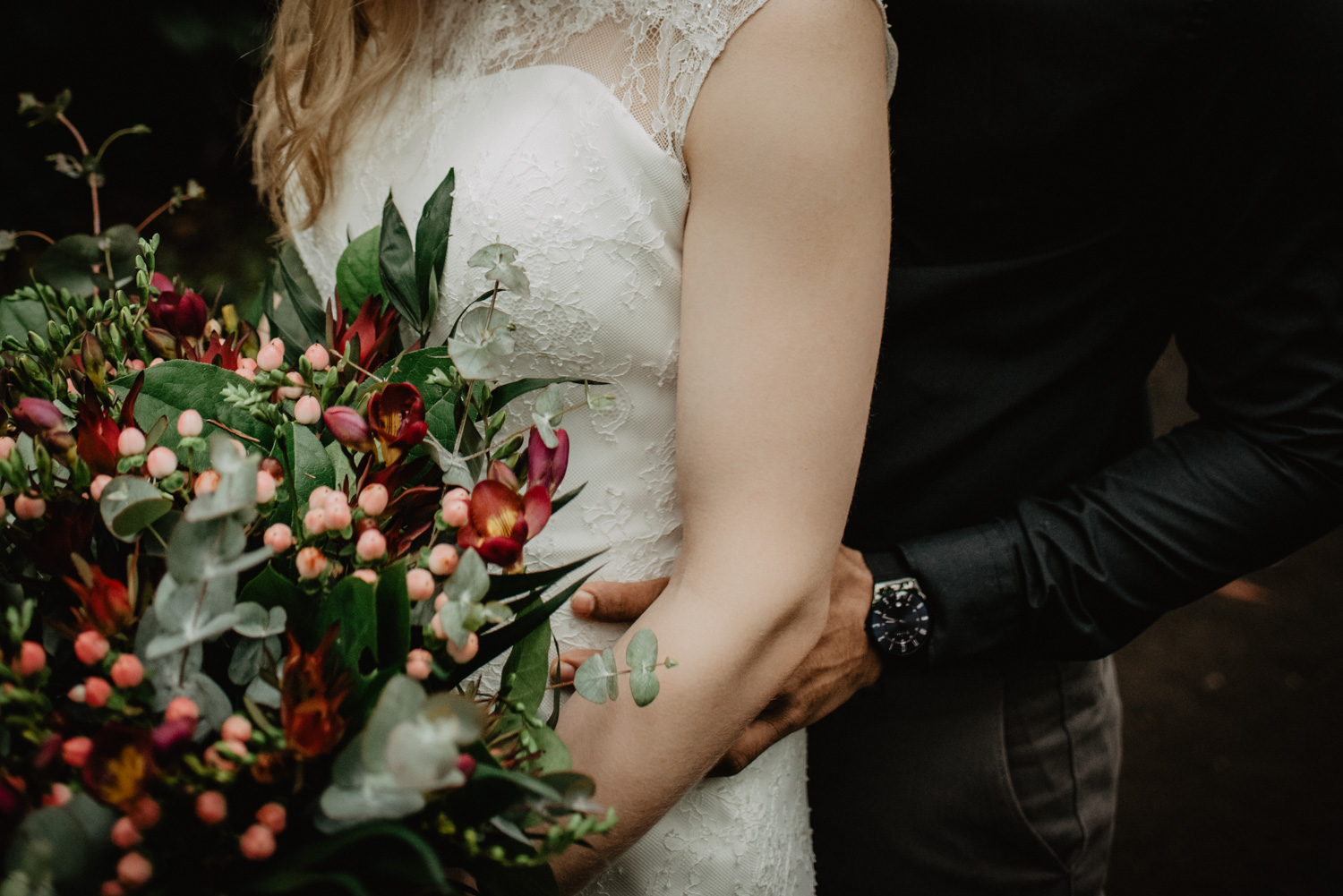 Ein Bräutigam umarmt seine Braut von hinten, während sie einen Boho-Blumenstrauß in den Händen hält.