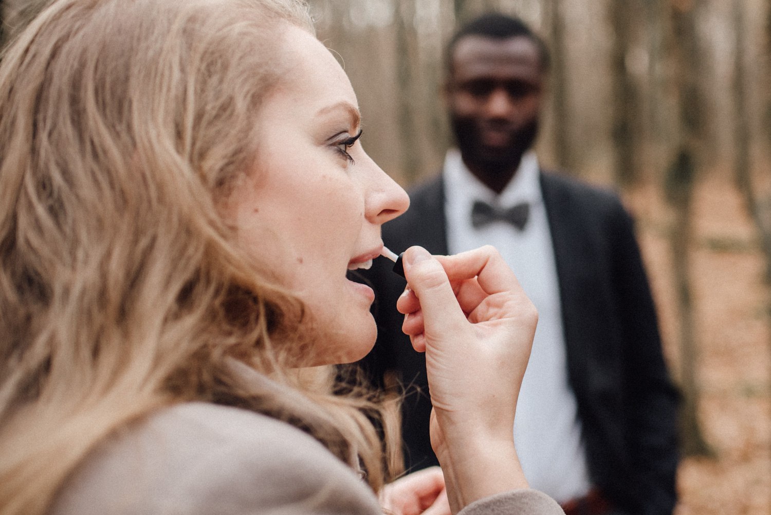 Eine Frau trägt Lipgloss auf, während ihr Mann sie im Hintergrund lächelnd beobachtet.